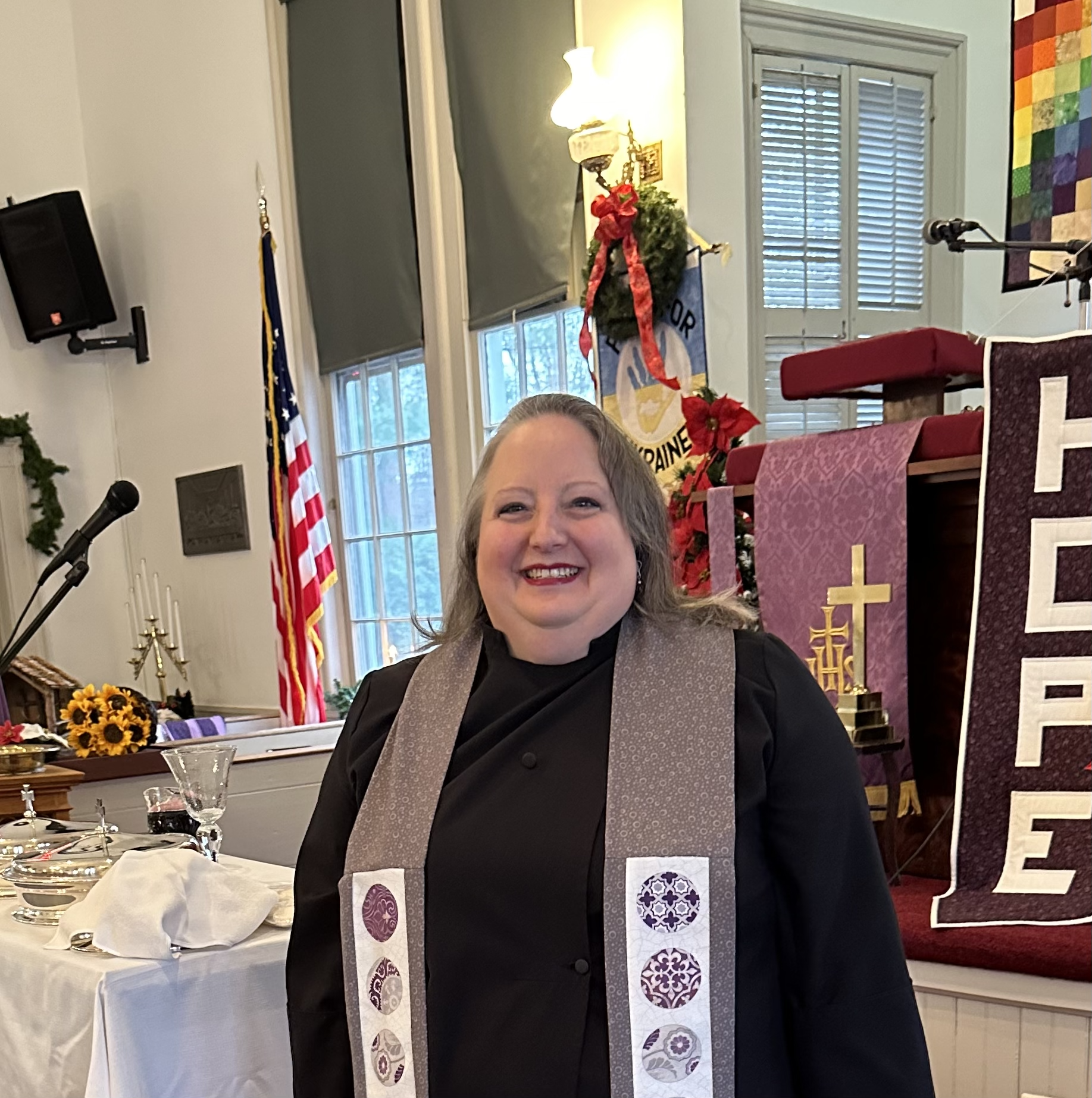 White woman with blond hair in a black clergy robe and purple and white stole standing in front of a pulpit with a purple cloth and a banner reading HOPE.